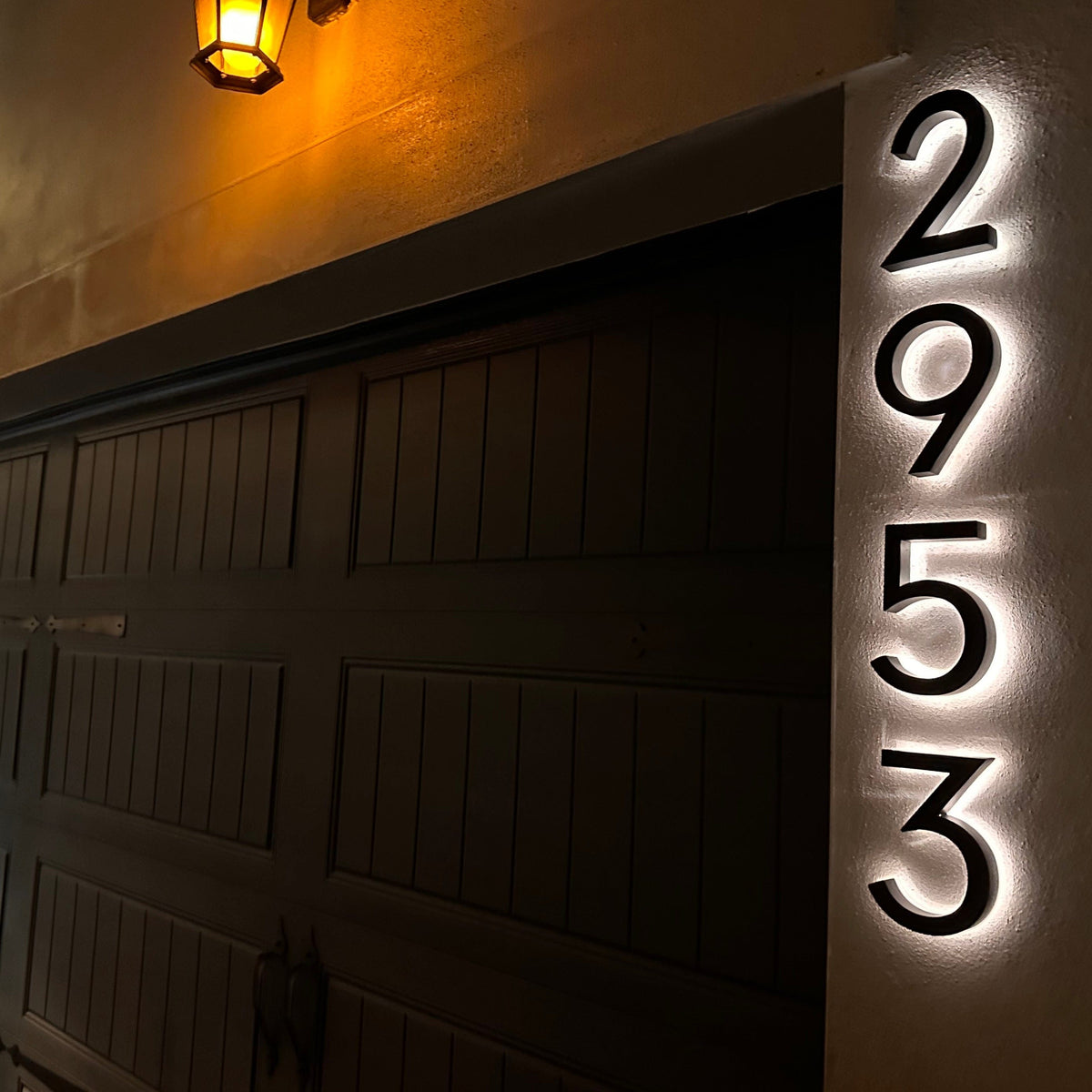 Vertical black backlit house numbers 2953 glowing white on a stucco garage pillar beside a lantern at night