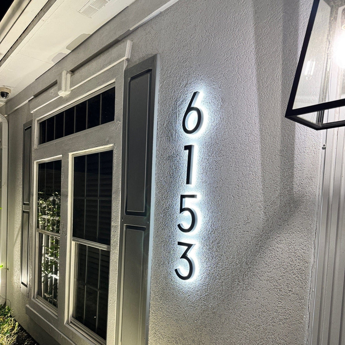 Vertical black backlit house numbers 6153 with a cool white glow on grey stucco beside a window at night