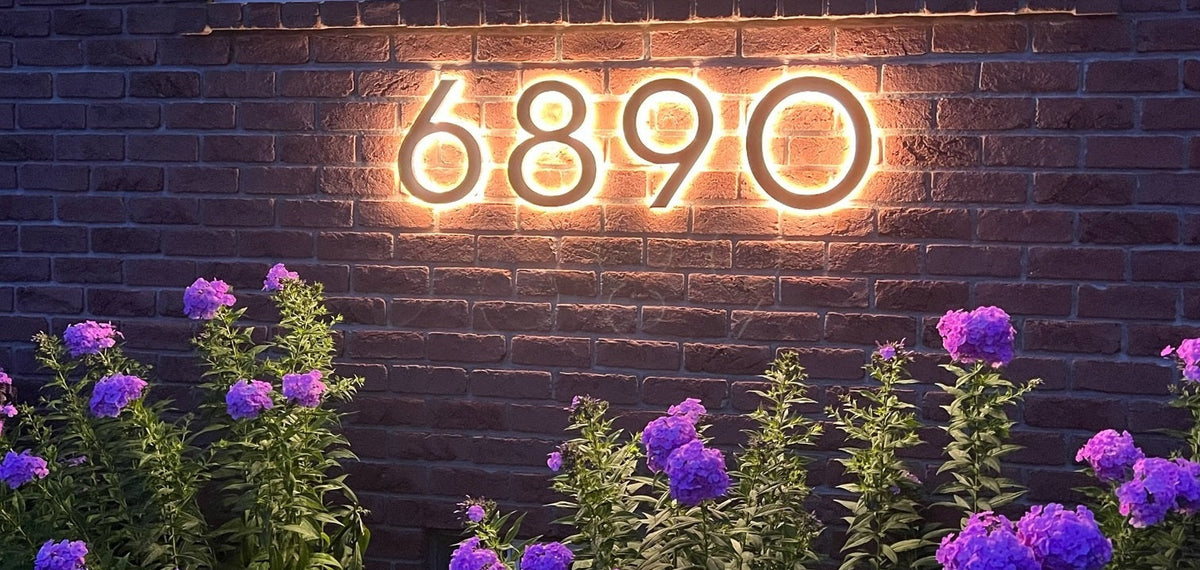 Backlit LED house numbers 6890 glowing warm white on a brick wall above purple flowers at night